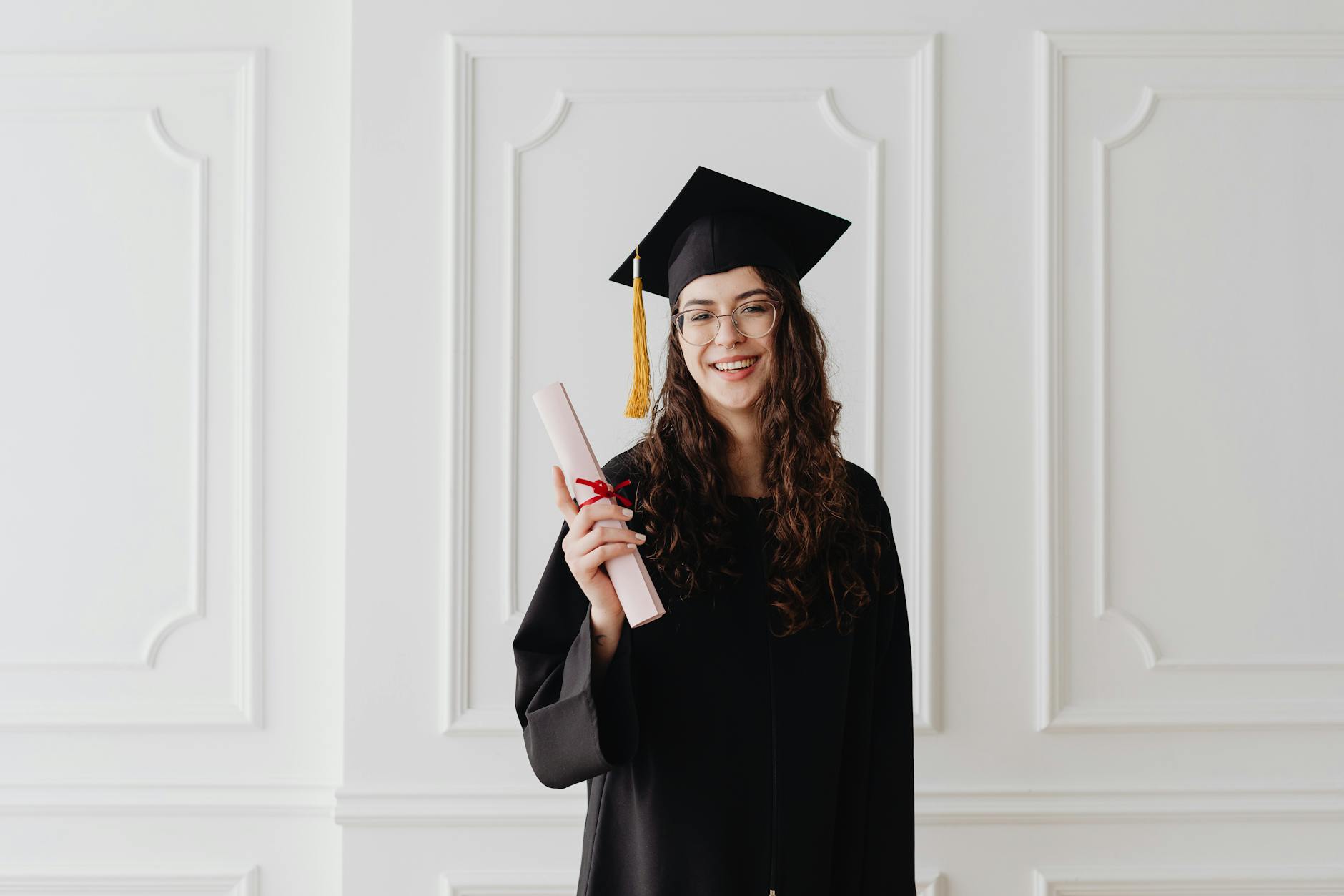 Smiling young woman in graduation gown and cap holding a diploma indoors. (Photo by www.kaboompics.com on Pexels)
